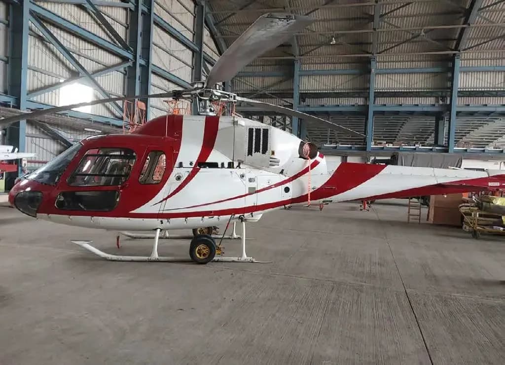 A red and white helicopter with a striped paint scheme parked inside a hangar, viewed from the left side.
