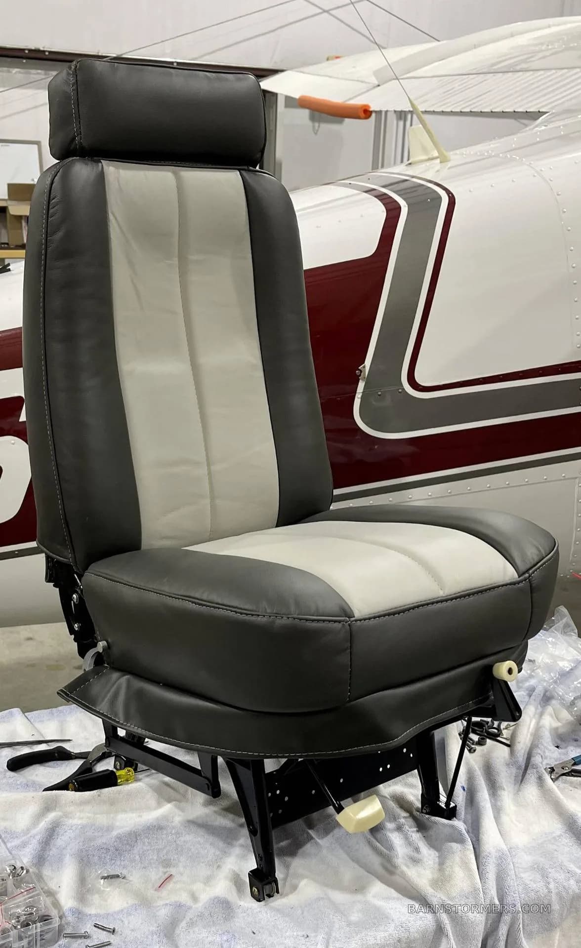 A newly upholstered gray and light gray aircraft seat is shown in front of the fuselage of a white and maroon aircraft.
