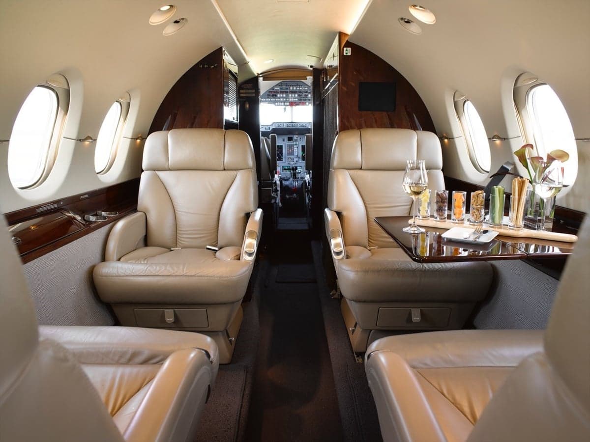 The private jet cabin shows tan leather seating and a table with refreshments, looking towards the cockpit.