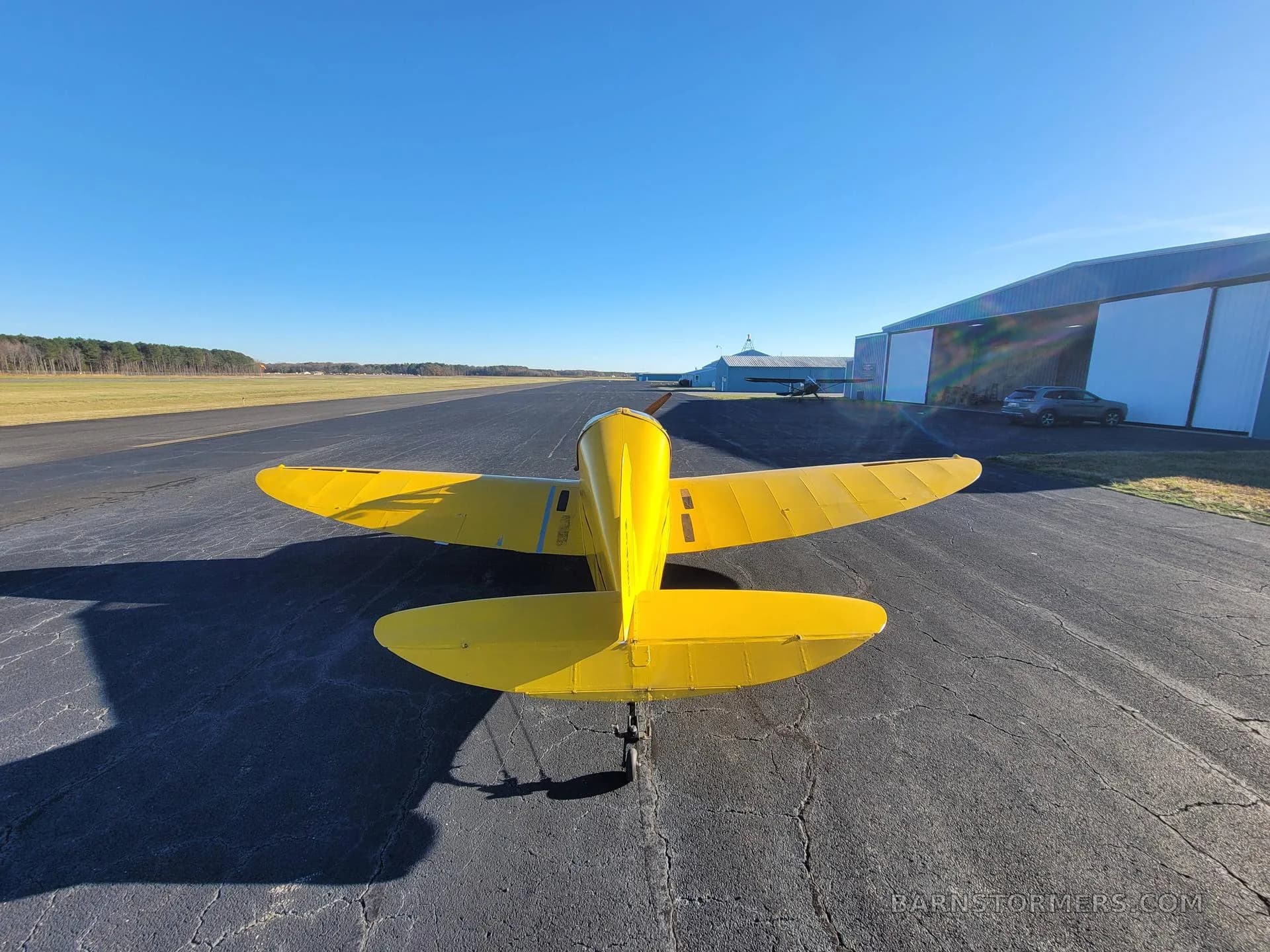 A rear view of a yellow single-engine aircraft with a tailwheel, parked on an asphalt runway with hangars in the background.