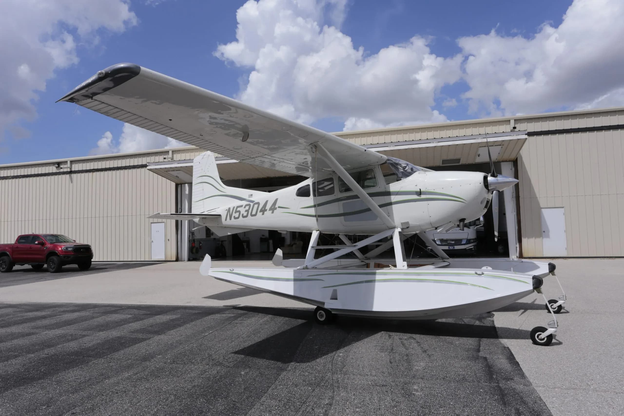 A front-side view of a white Cessna 180 Skywagon seaplane with green stripes, registration N53044, parked outside a hangar with a red pickup truck nearby.