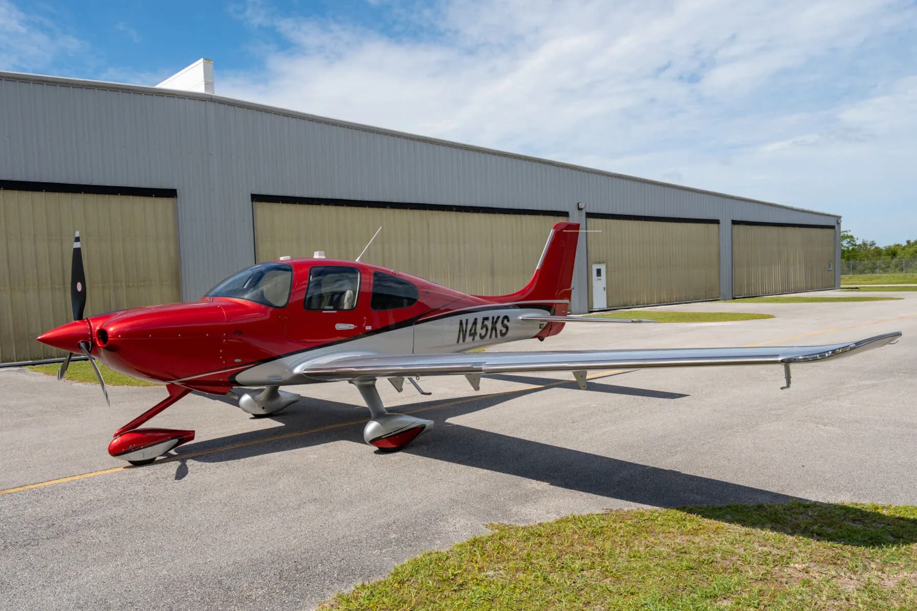A red and silver Cirrus SR22 GTS (N45KS) parked on an airfield with a hangar in the background.