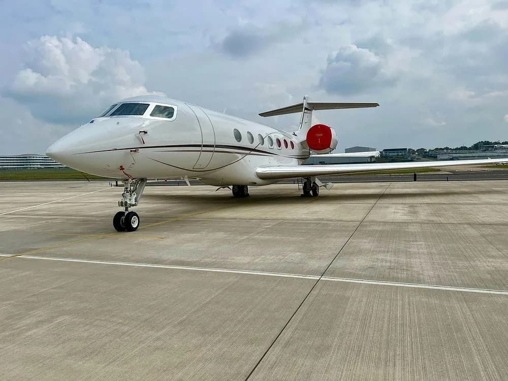 A white Gulfstream G650 business jet with red engine nacelles parked on a concrete tarmac under a cloudy sky.