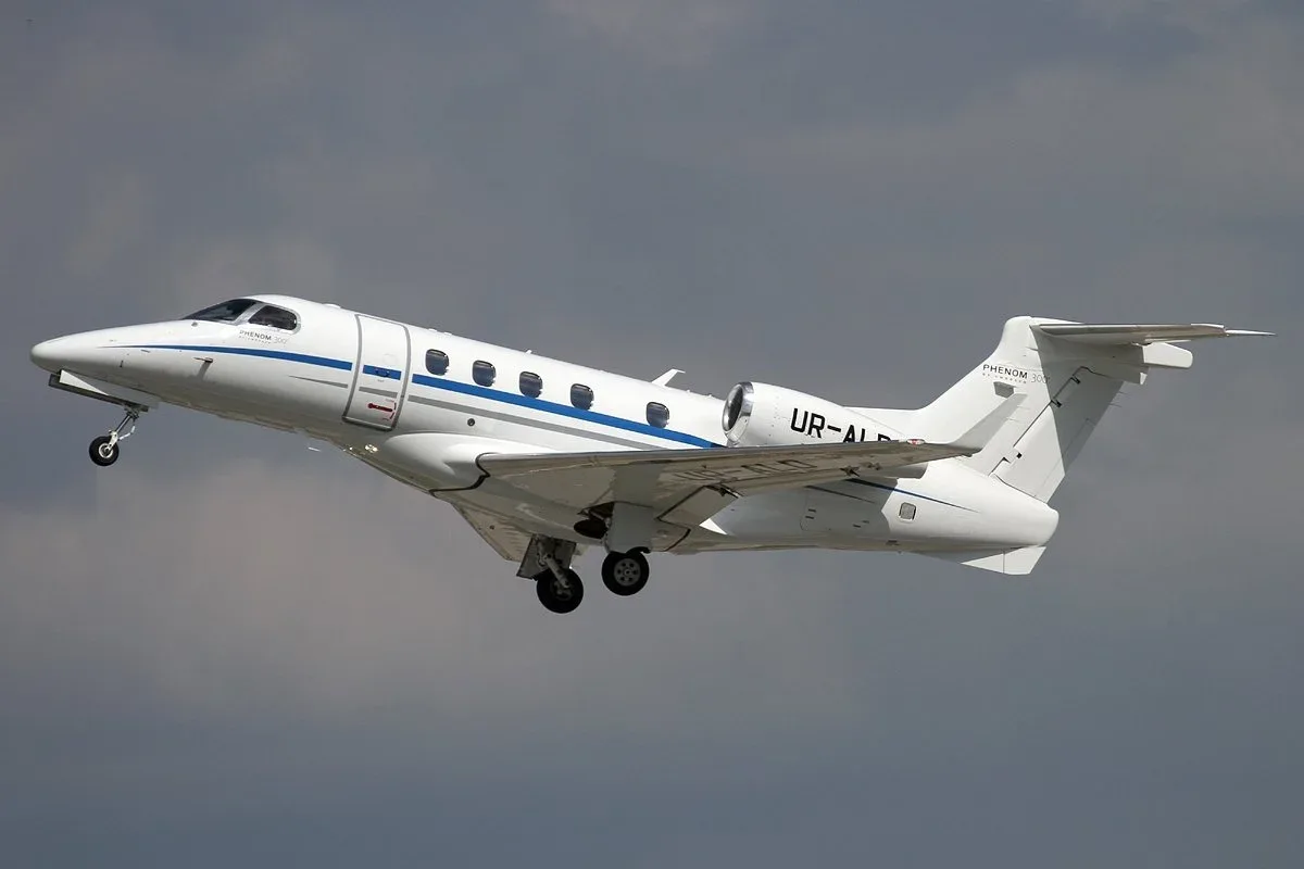 A Phenom 300 aircraft with registration UR-ALD is shown in flight with a white and blue paint scheme.