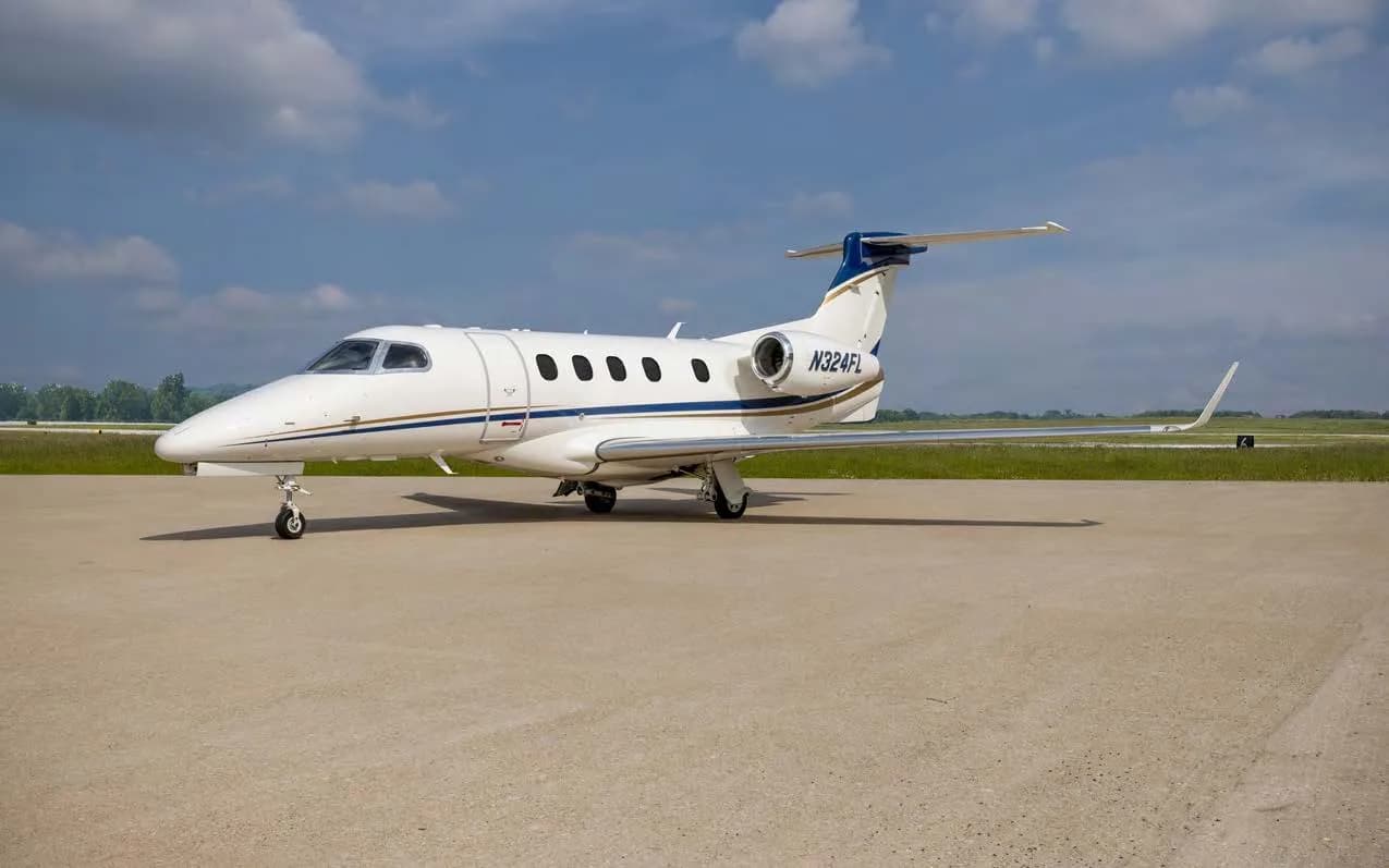 A white Embraer Phenom 300 private jet with blue and gold stripes on the tail and fuselage, parked on an airfield under a partly cloudy sky.