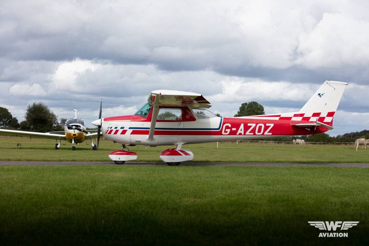 A red and white Cessna 150 Aerobat (G-AZOZ) with a checkered tail, parked on a grassy airfield next to another aircraft.