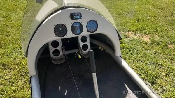 The cockpit of a white ultralight aircraft shows the instrument panel with gauges and foot pedals.