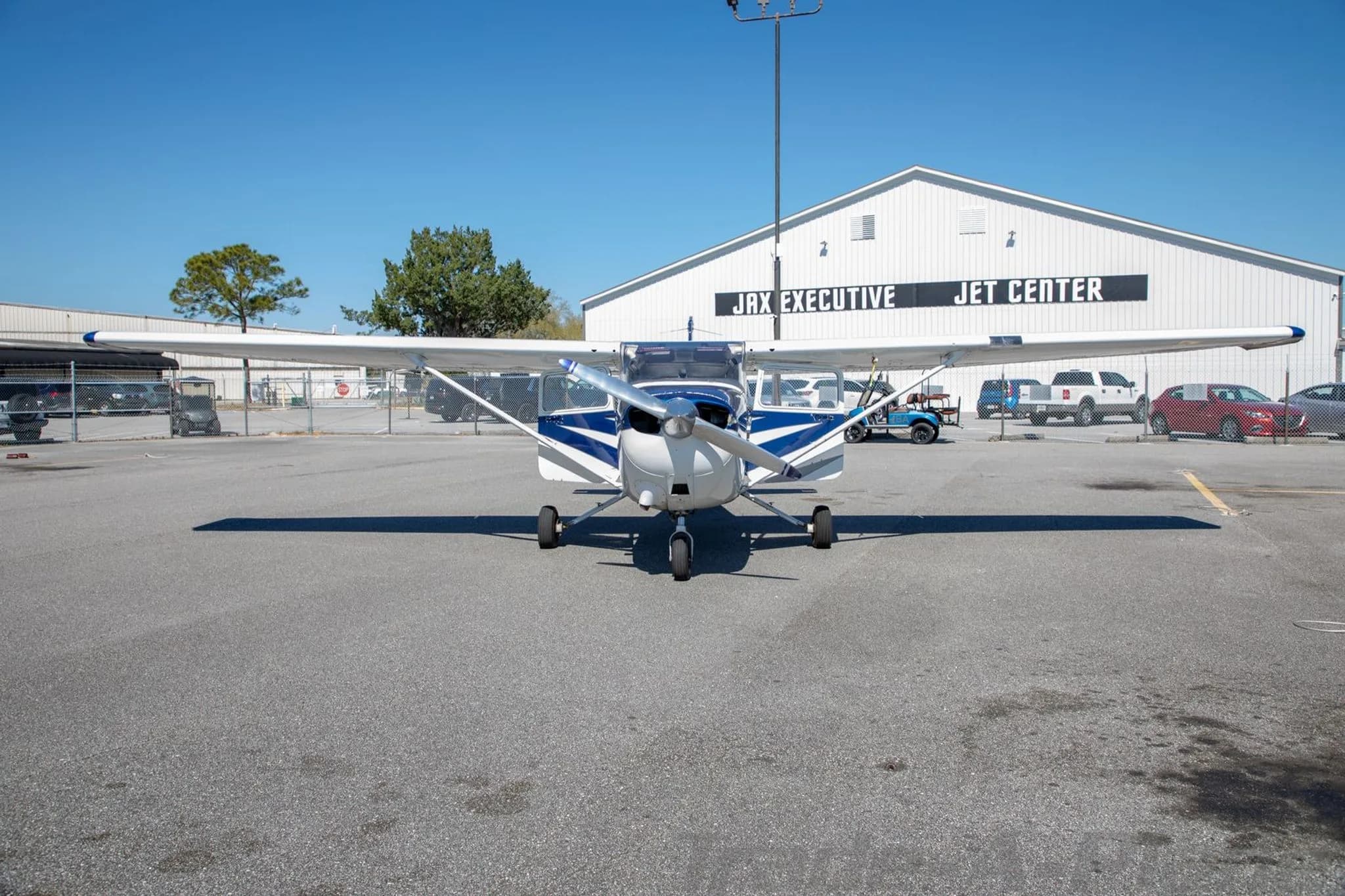 A wide front view of the N86ER aircraft, a white and blue Cessna 172, parked on an airfield with a large white hangar in the background.