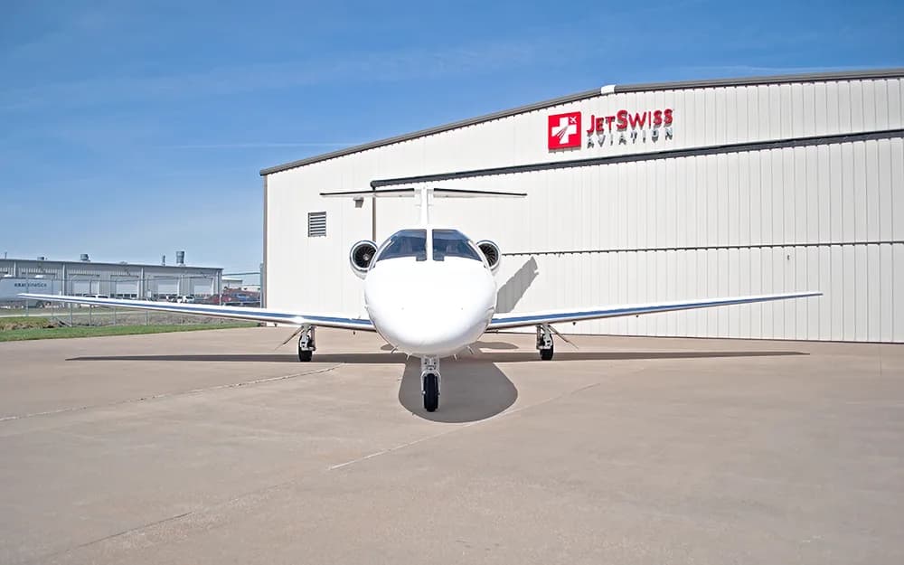 A white business jet with a blue stripe on the fuselage, parked head-on in front of a JetSwiss Aviation hangar.