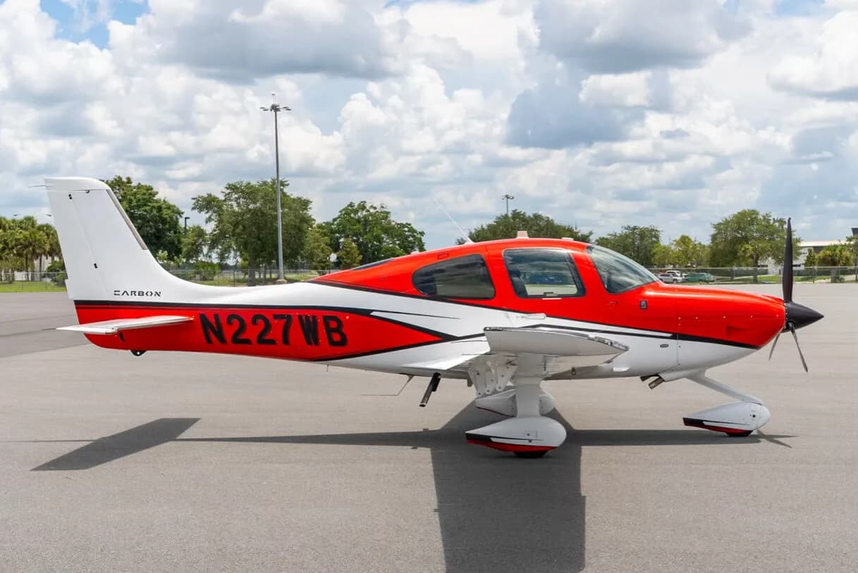 A red and white Cirrus SR22 aircraft with registration N227WB parked on an airfield under a partly cloudy sky.