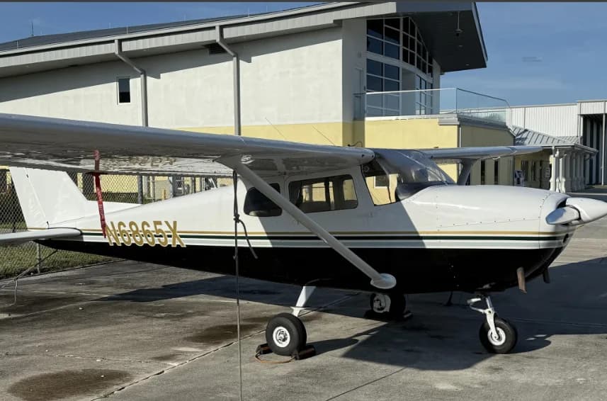 A side view of a white and black Cessna 172 with registration N6865X, parked on an airfield in front of a building.