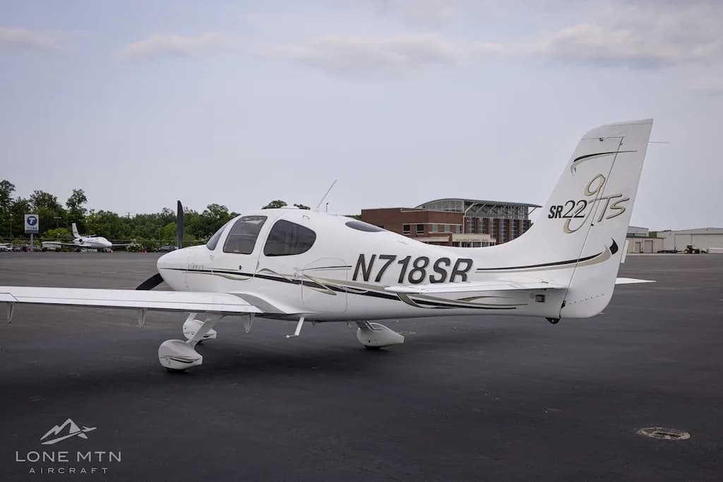 Rear-side view of a white Cirrus SR22T aircraft with black and gold accents parked on an asphalt tarmac with buildings in the background.