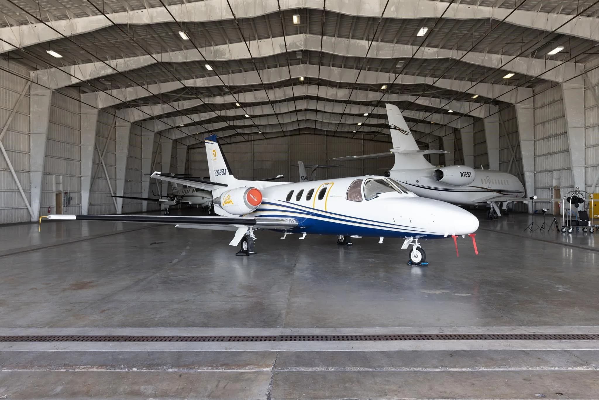 A white and blue Cessna Citation jet (N3095M) parked inside a large hangar with another jet in the background.