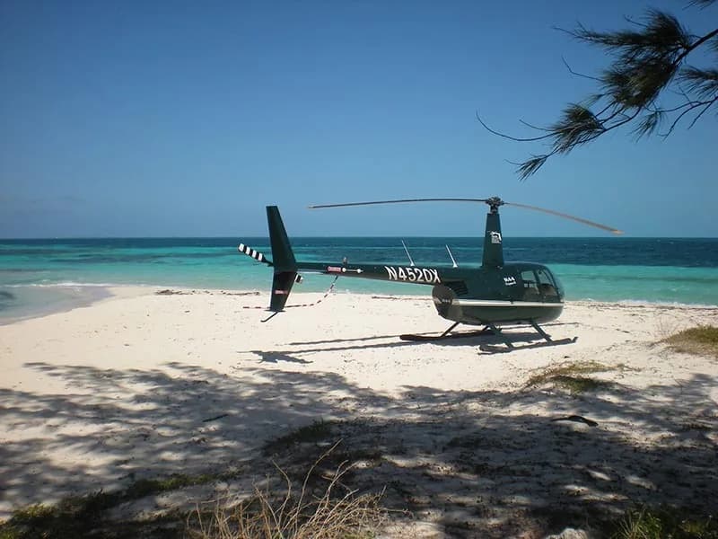 A dark green Robinson R44 Clipper II helicopter, registration N4520X, is parked on a sunny white sand beach with turquoise water in the background.