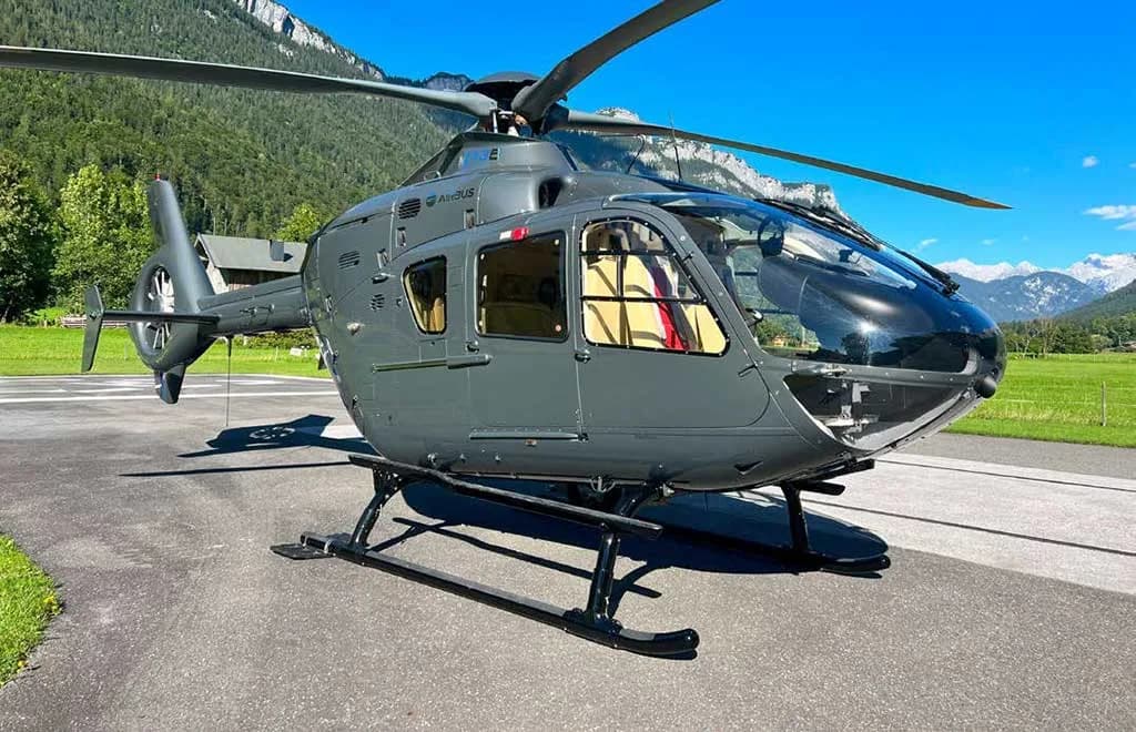 A front-side view of a grey Airbus H135 helicopter parked on an asphalt pad.