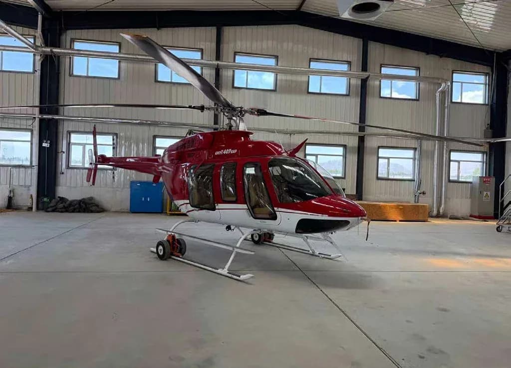 A red and white Bell 407GXP helicopter is parked inside a hangar with many windows.