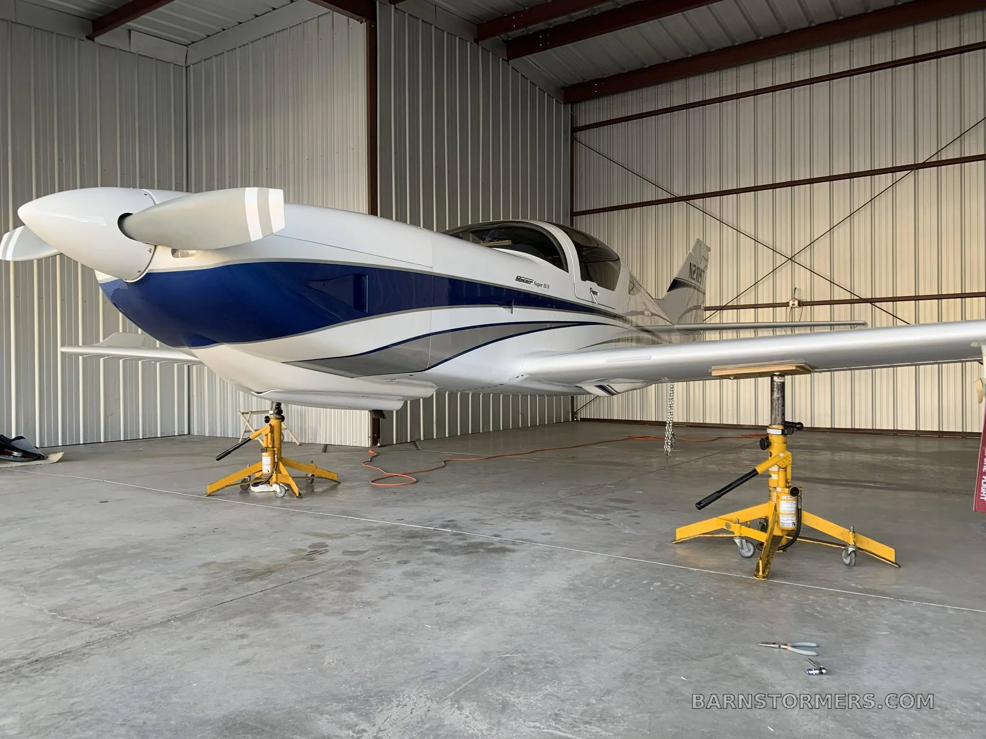 A white and blue single-engine aircraft (N210ST) is shown from a front-left view on maintenance jacks inside a hangar.