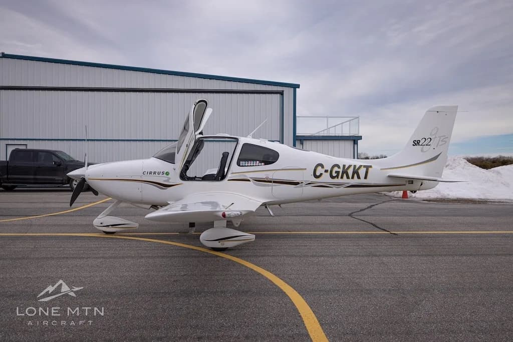 A side view of a white Cirrus SR22 aircraft with registration C-GKKT, doors open, parked on an airfield.