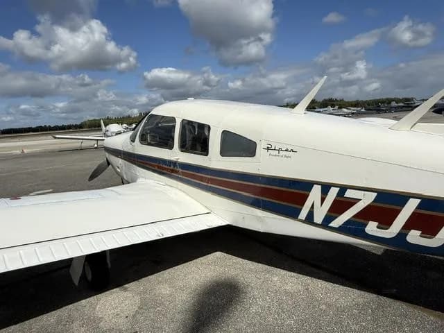 A close-up side view of the white Piper Cherokee aircraft N7JJ, showing its registration and "Piper Freedom of Flight" decal.