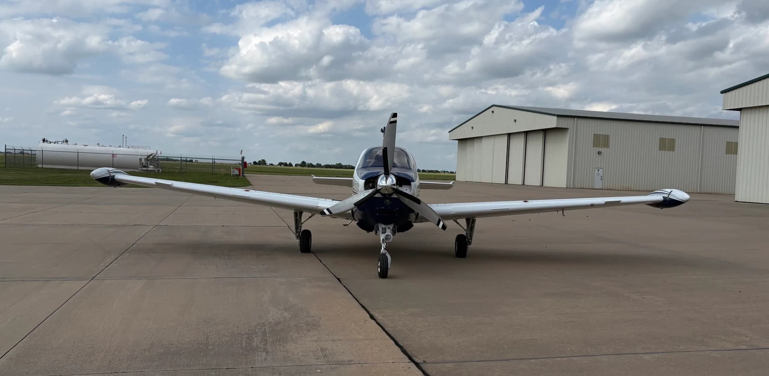 A head-on view of the white and blue Beechcraft Bonanza N686AG parked on an airfield tarmac under a cloudy sky.