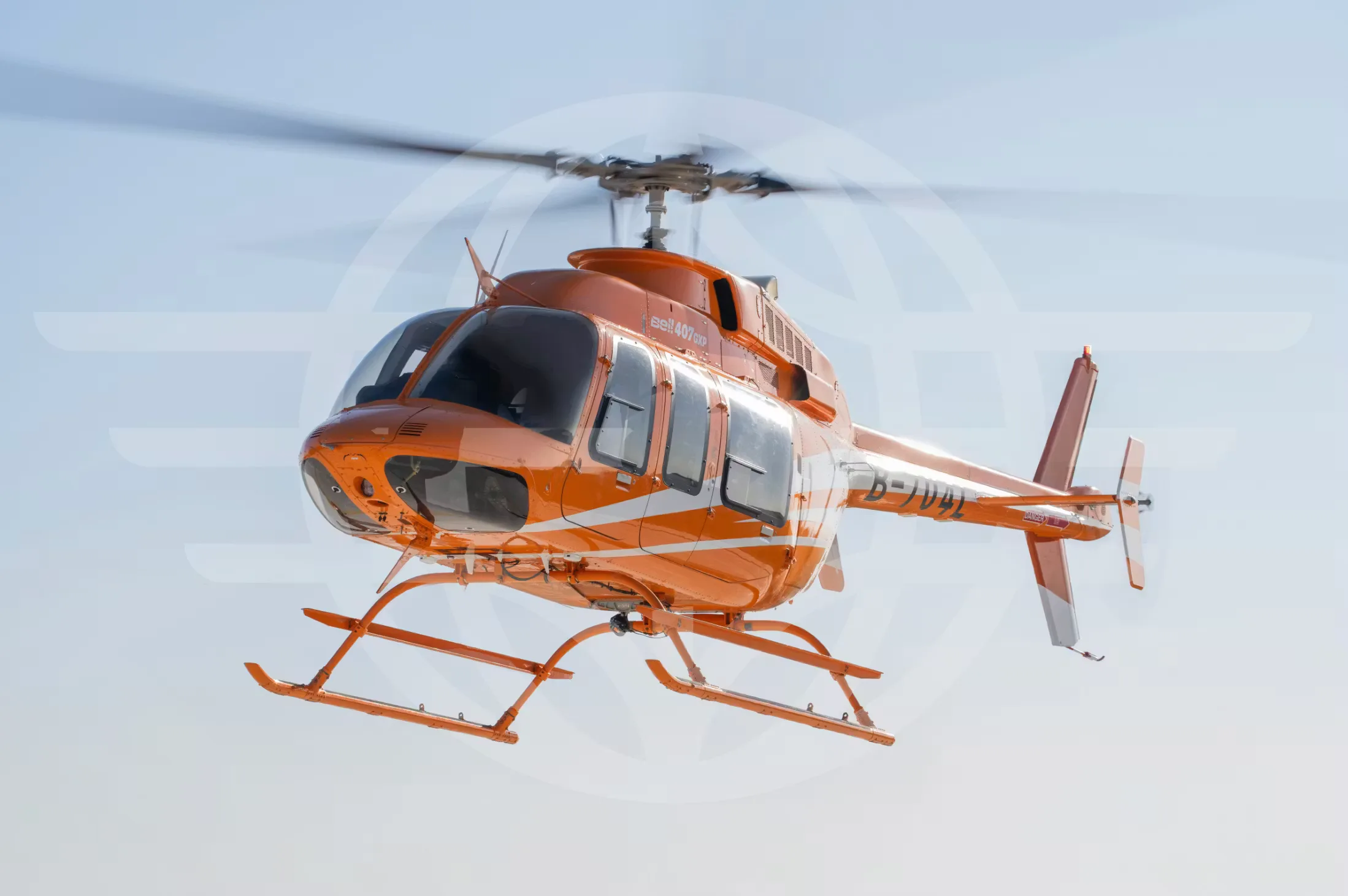 An orange and white Bell 407GXi helicopter is in flight against a light blue sky.