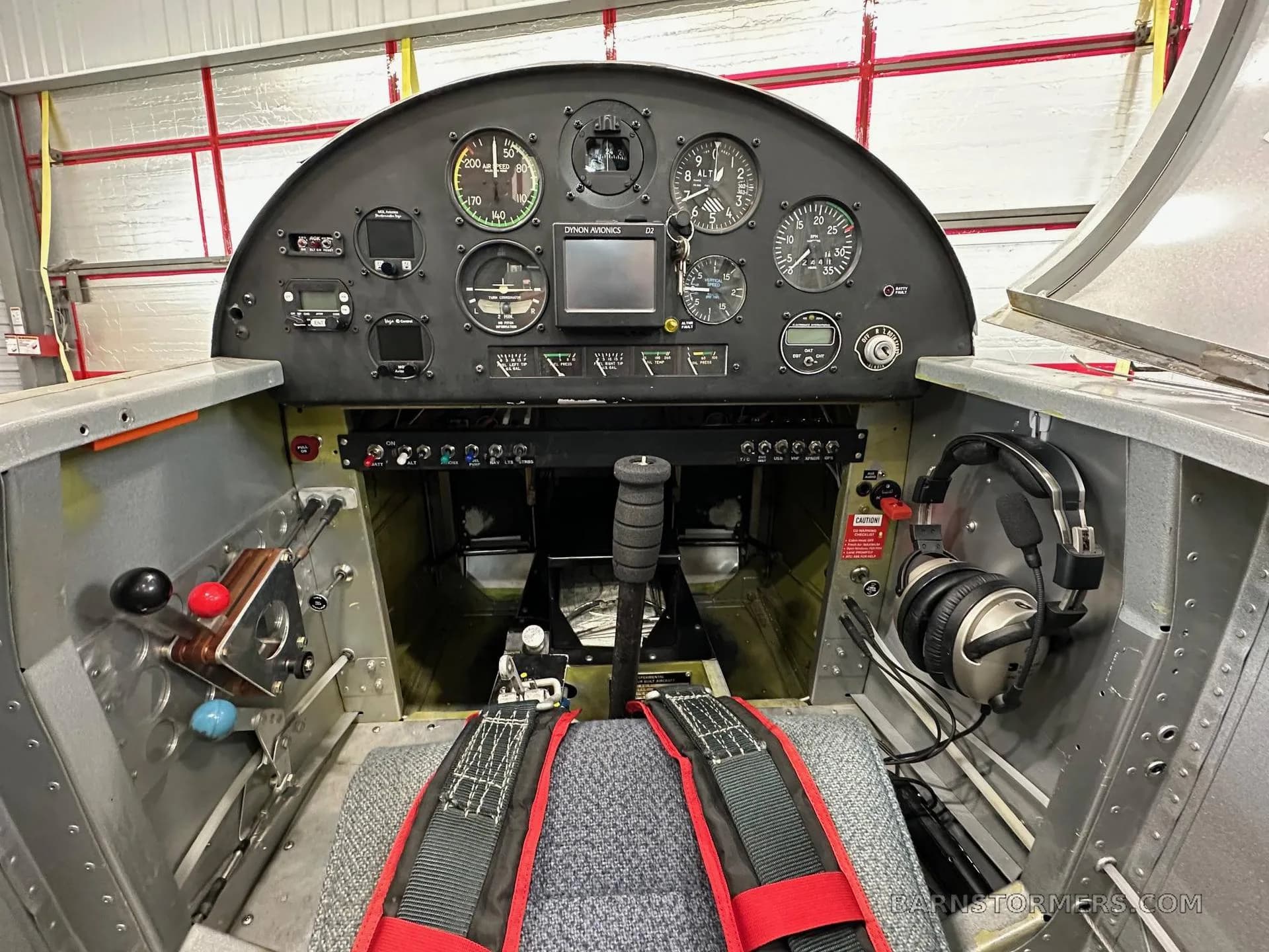 The cockpit of a single-seat aircraft, showing the instrument panel, control stick, and seat with red harnesses.