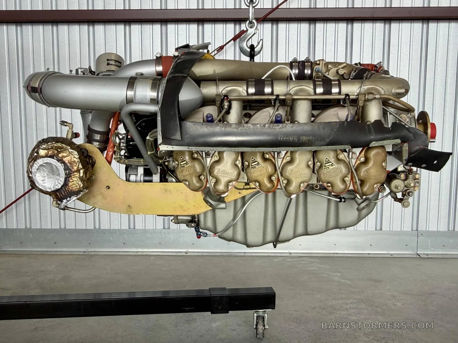 A side view of a Continental aircraft engine, showing the exhaust and ignition components, suspended in a hangar.