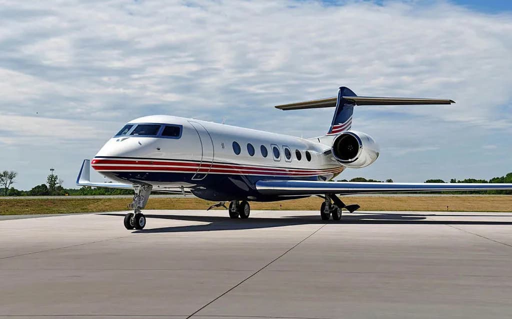 A white business jet with blue and red stripes parked on an airport tarmac under a partly cloudy sky.