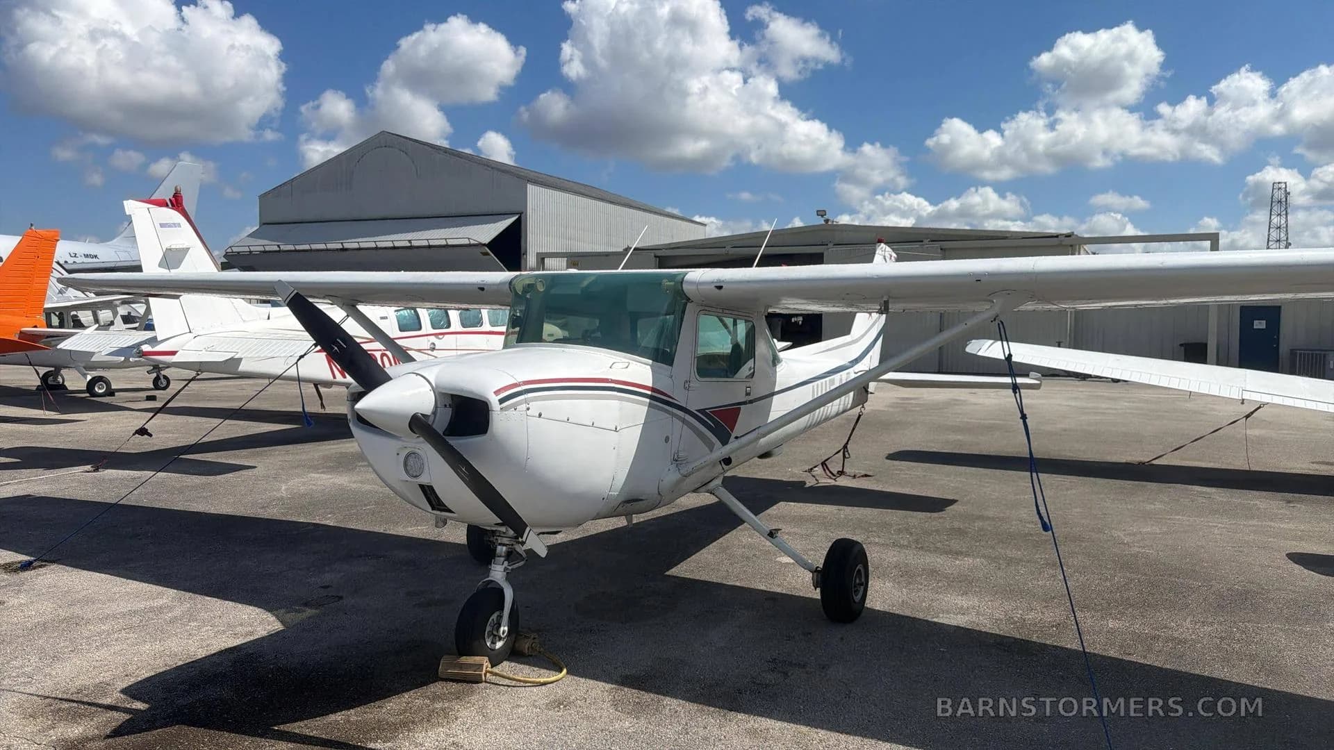 A white Cessna 172 with red and black stripes parked on an airport tarmac under a cloudy sky.