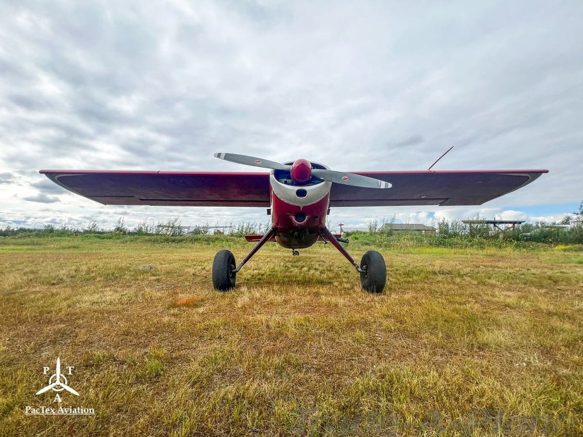 A head-on view of a red and silver single-engine aircraft with a propeller and large tires, parked on a grassy field under a cloudy sky.