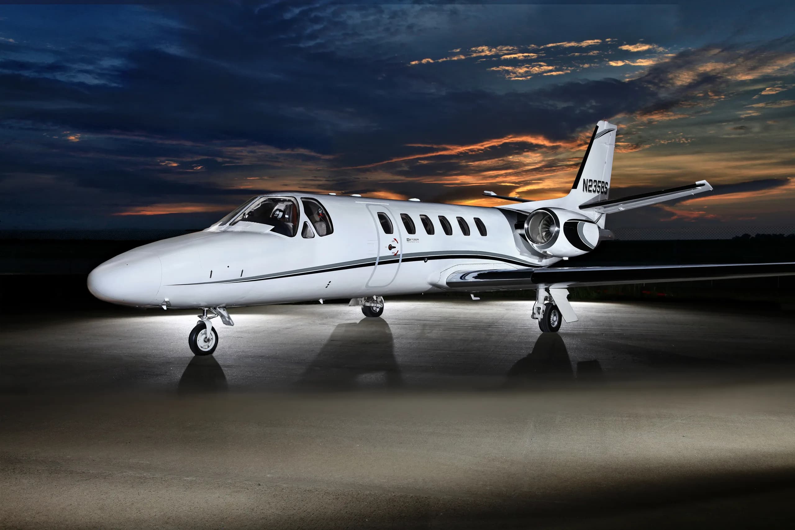 A white business jet with black and grey stripes, registration N235BS, parked on an tarmac at dusk with a dramatic sky.