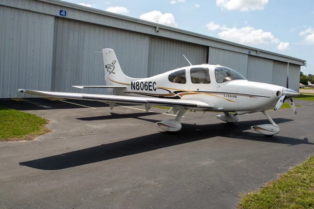 A white Cirrus SR22 aircraft with black and gold stripes, registration N806EC, parked on an asphalt tarmac in front of a hangar.