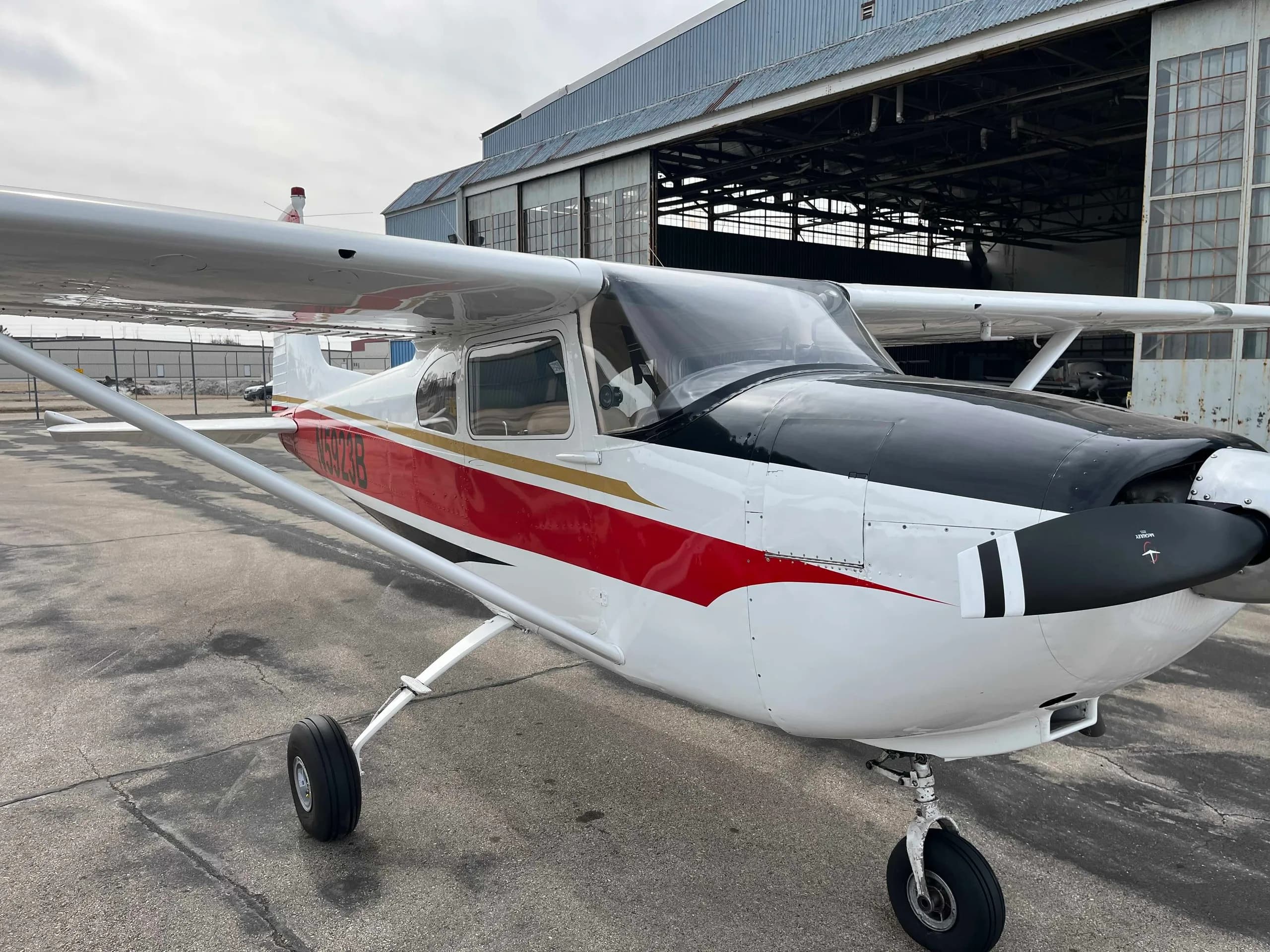 A white Cessna 182 aircraft with red and black stripes on the fuselage, parked on an asphalt apron with a hangar in the background.