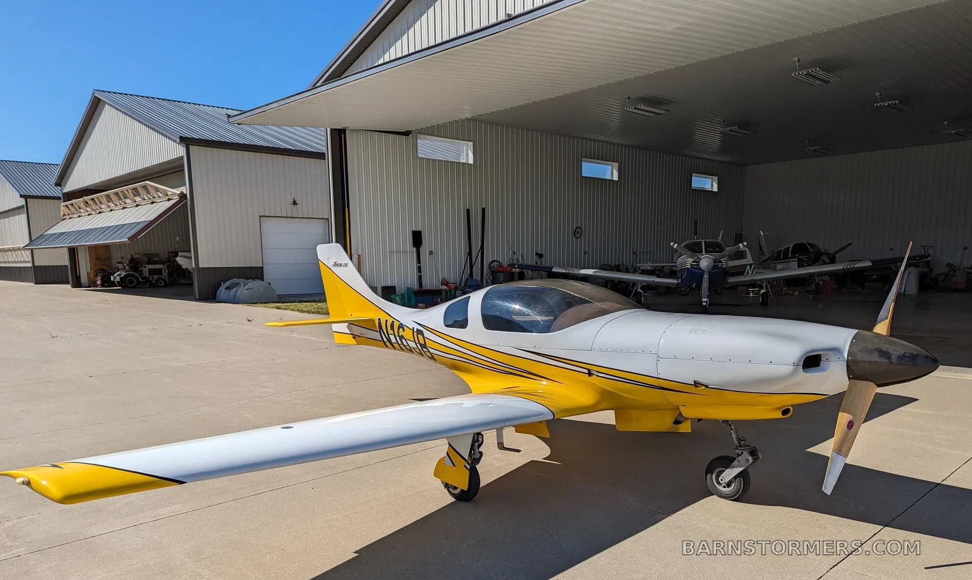 A white and yellow single-engine aircraft with a propeller, parked on concrete outside an open hangar.