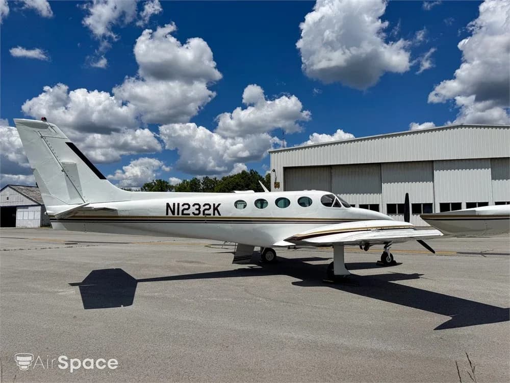 A white twin-engine aircraft with gold and brown stripes parked on an asphalt ramp next to a hangar.