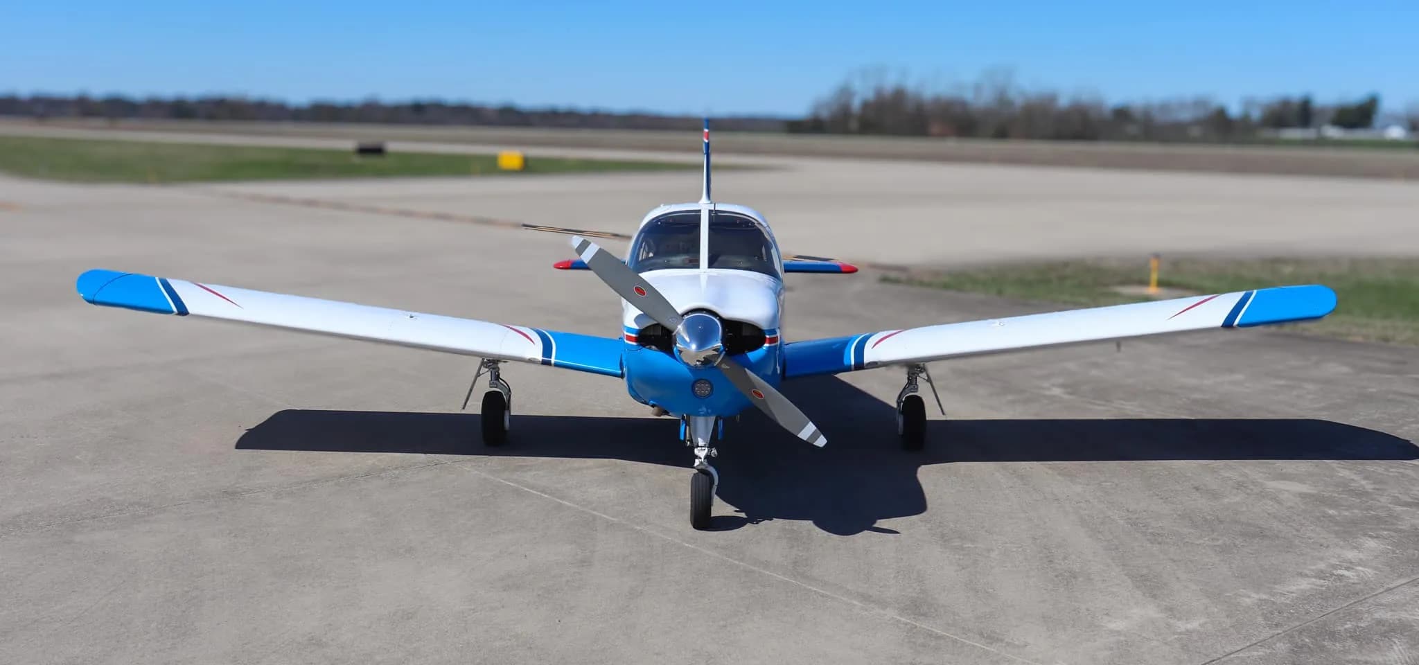A head-on view of a blue and white Piper Cherokee aircraft with red stripes parked on an airfield.