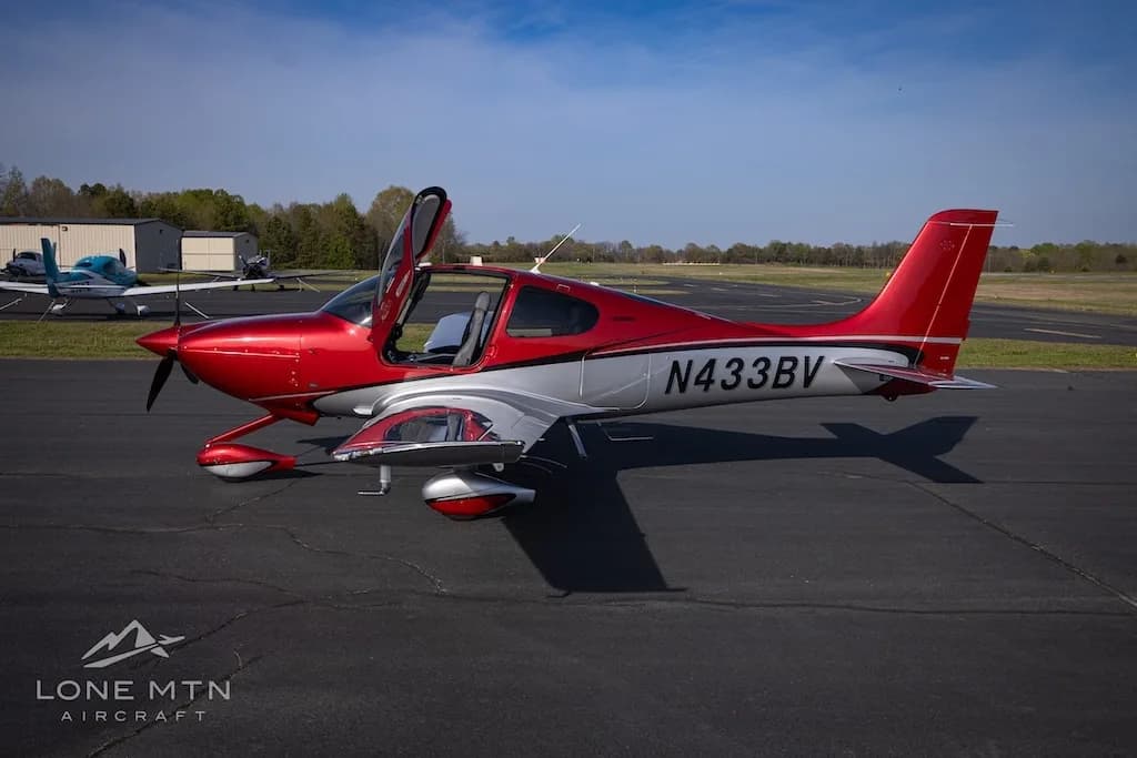 A side view of the red and silver Cirrus SR22 aircraft N433BV with its doors open, parked on an airfield.