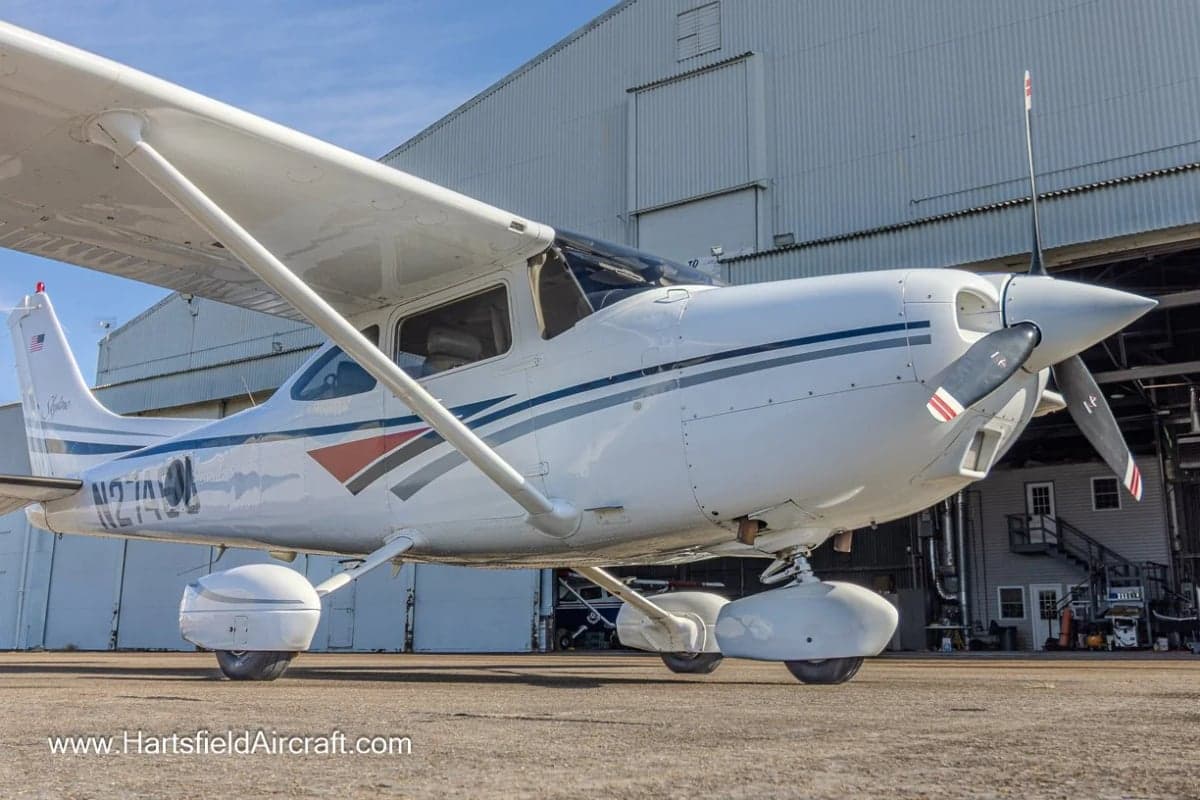 A Cessna Skylane aircraft with a white and gray paint scheme parked in front of a hangar.