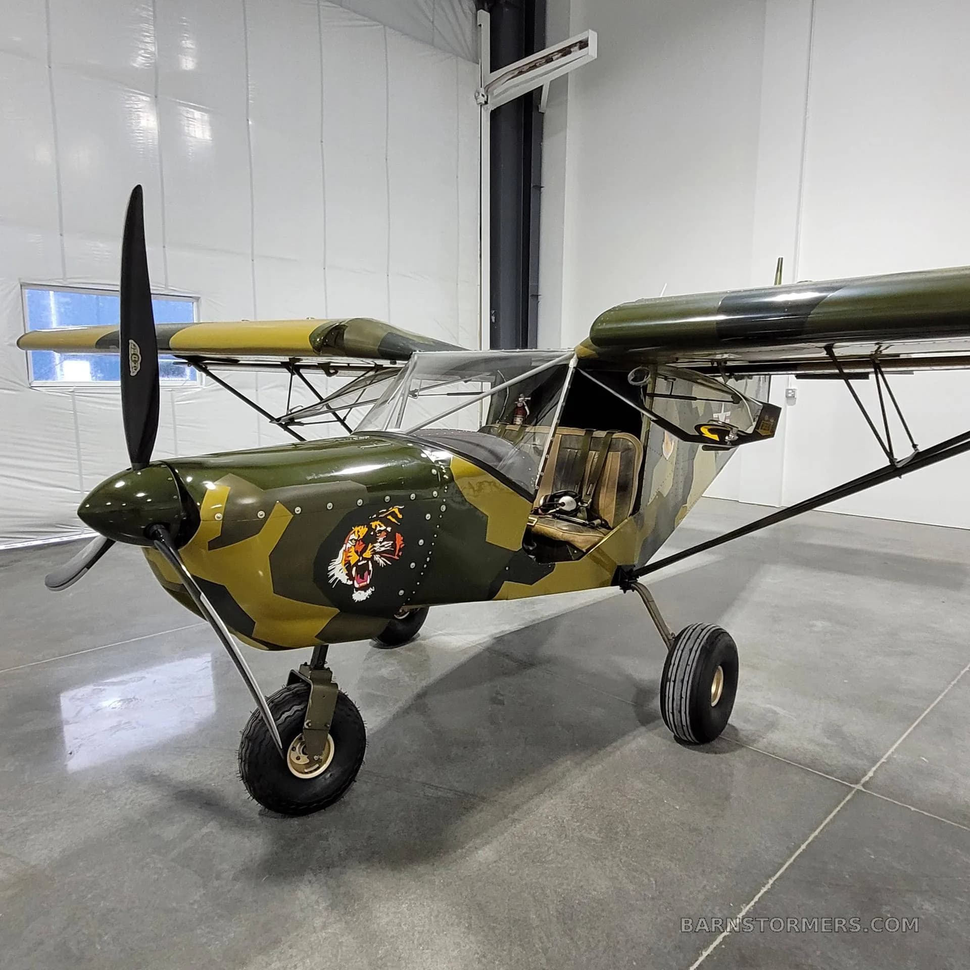 A small, single-engine, high-wing aircraft with a green and brown camouflage paint scheme and a tiger head decal on the nose, parked in a hangar.