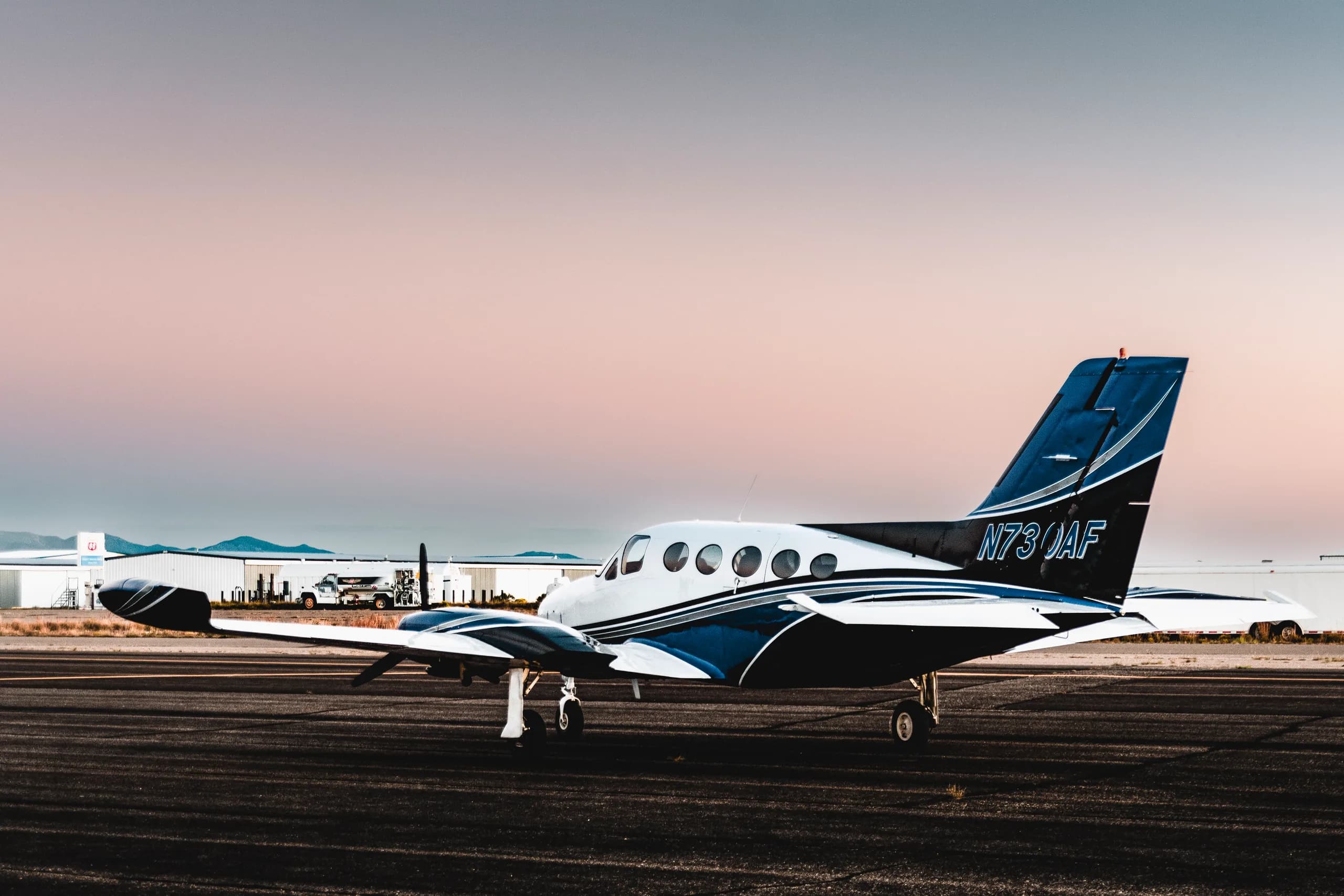 A white and blue twin-engine aircraft with registration N730AF on the tail, parked on a runway at sunset.