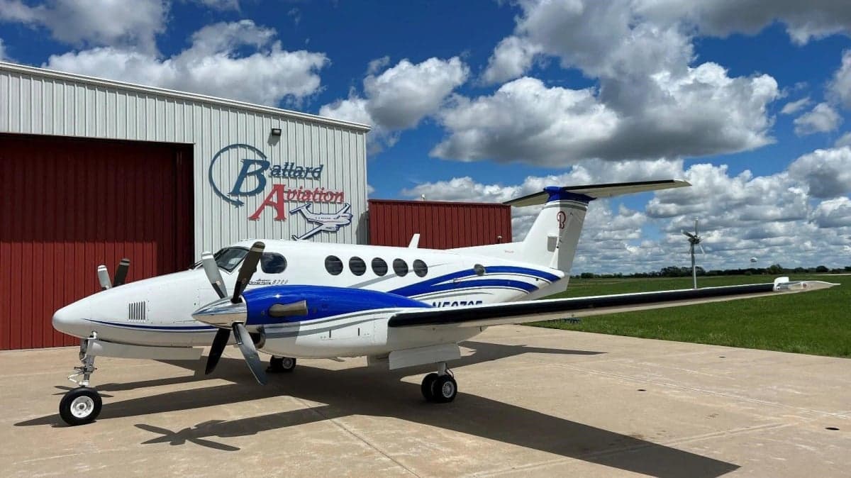 A Beechcraft King Air sits on the tarmac in front of a hangar with blue and white paint scheme.