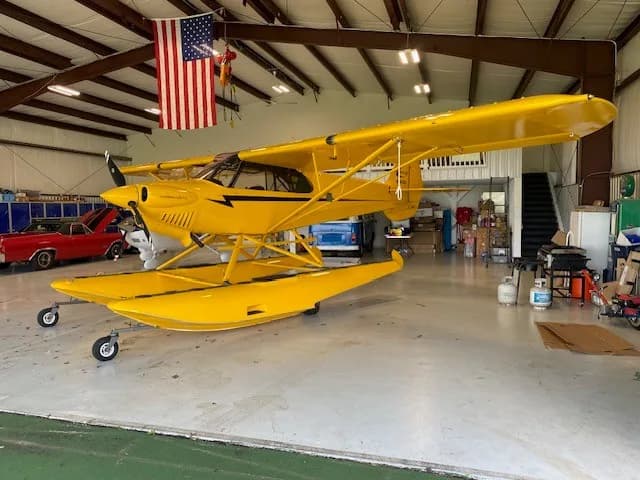 Side view of a yellow Carbon Cub floatplane with black stripes, parked inside a hangar.