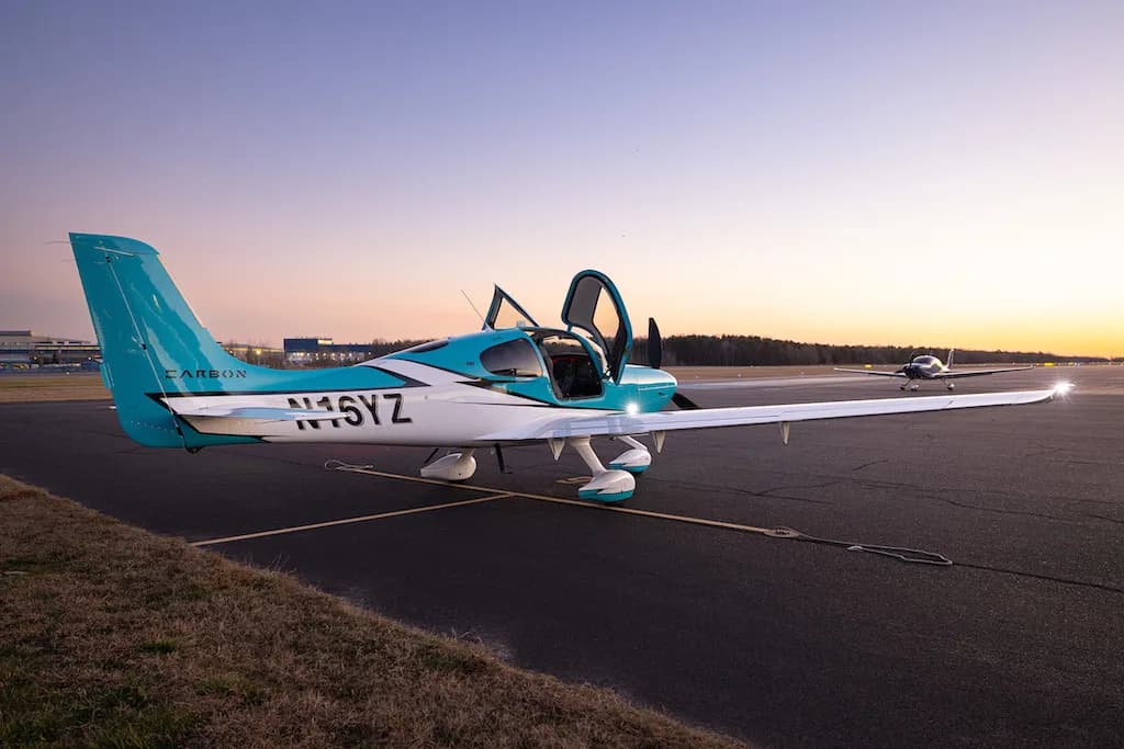 A turquoise and white Cirrus SR22 aircraft with its doors open, parked on an airfield with another plane in the distance at dusk.