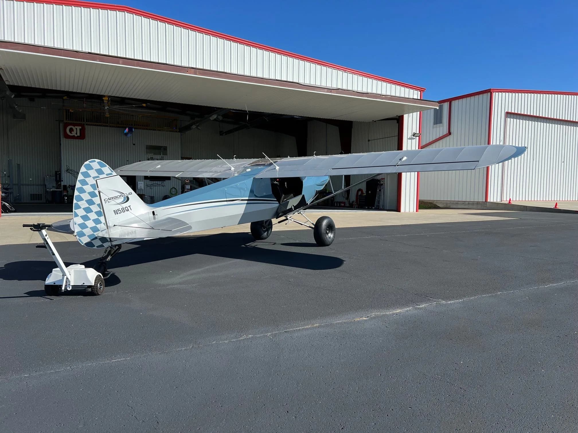 A rear-side view of a blue and grey Carbon Cub FX-3 aircraft with registration N58QT being moved by a tug outside a hangar.