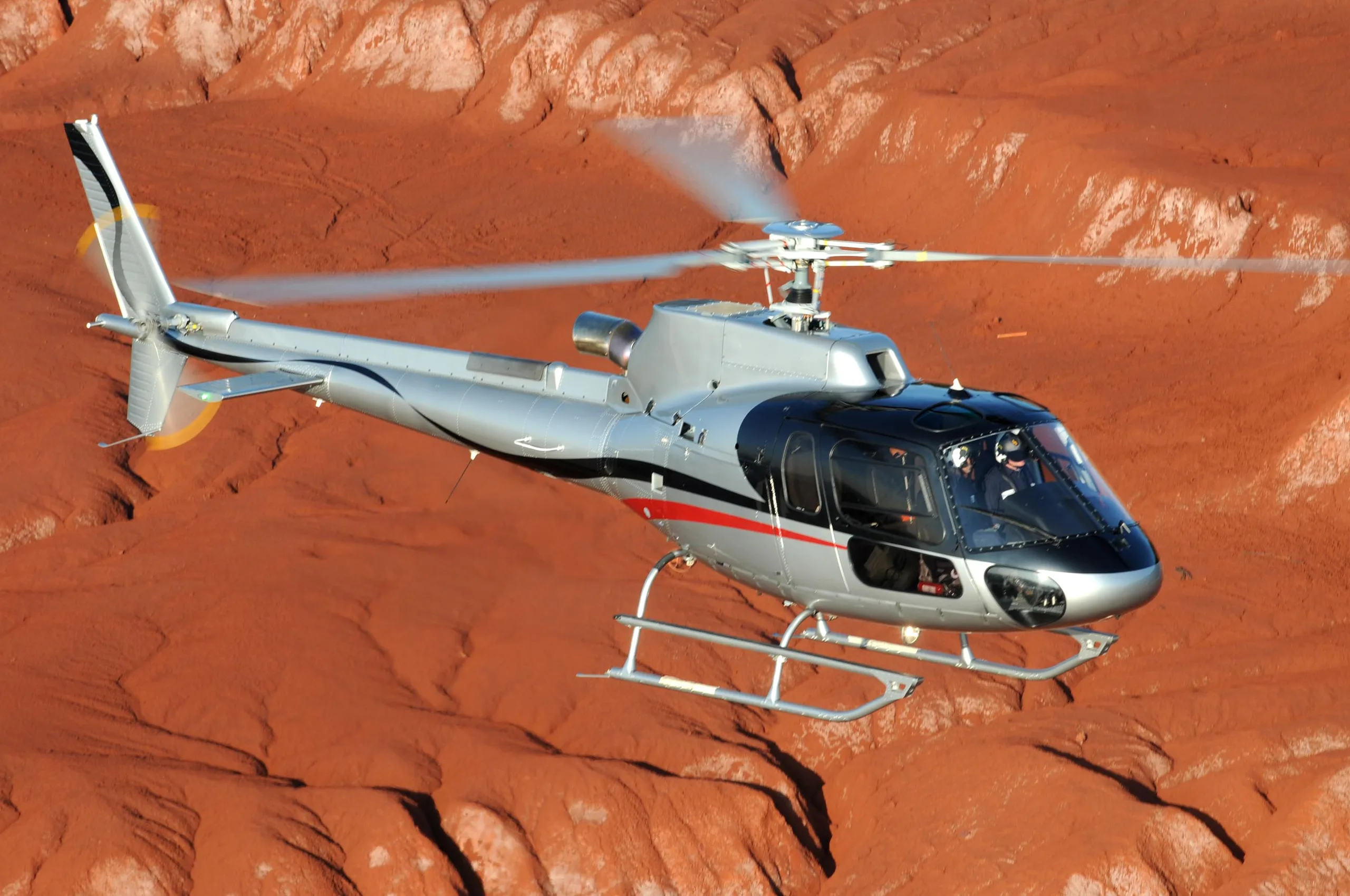 A silver and black helicopter flies over a red rock landscape.