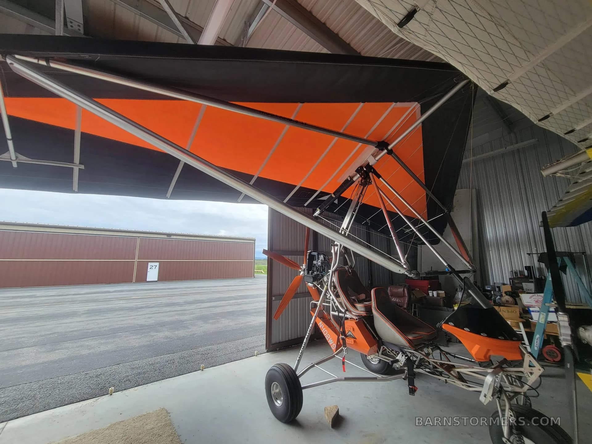 An orange and black weight-shift control ultralight aircraft parked inside a hangar with the door open to an airfield.