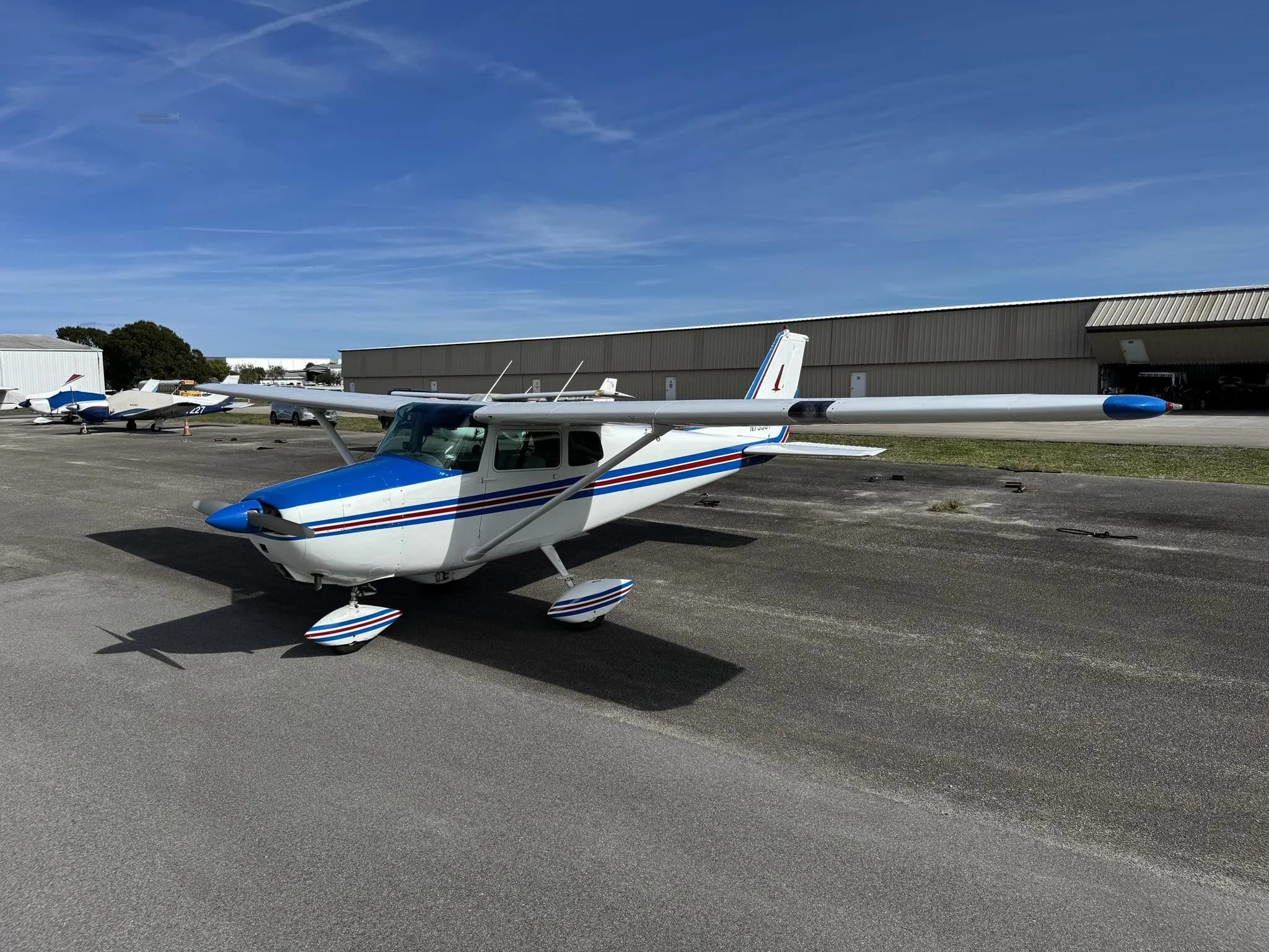 A white and blue Cessna 172 aircraft parked on an asphalt apron with hangars in the background.