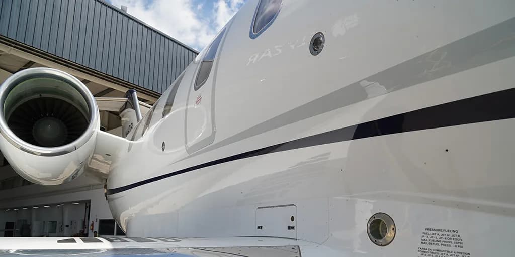A close-up view of the white fuselage and engine of a business jet, showing fuel port details and a dark blue stripe, parked near a hangar.