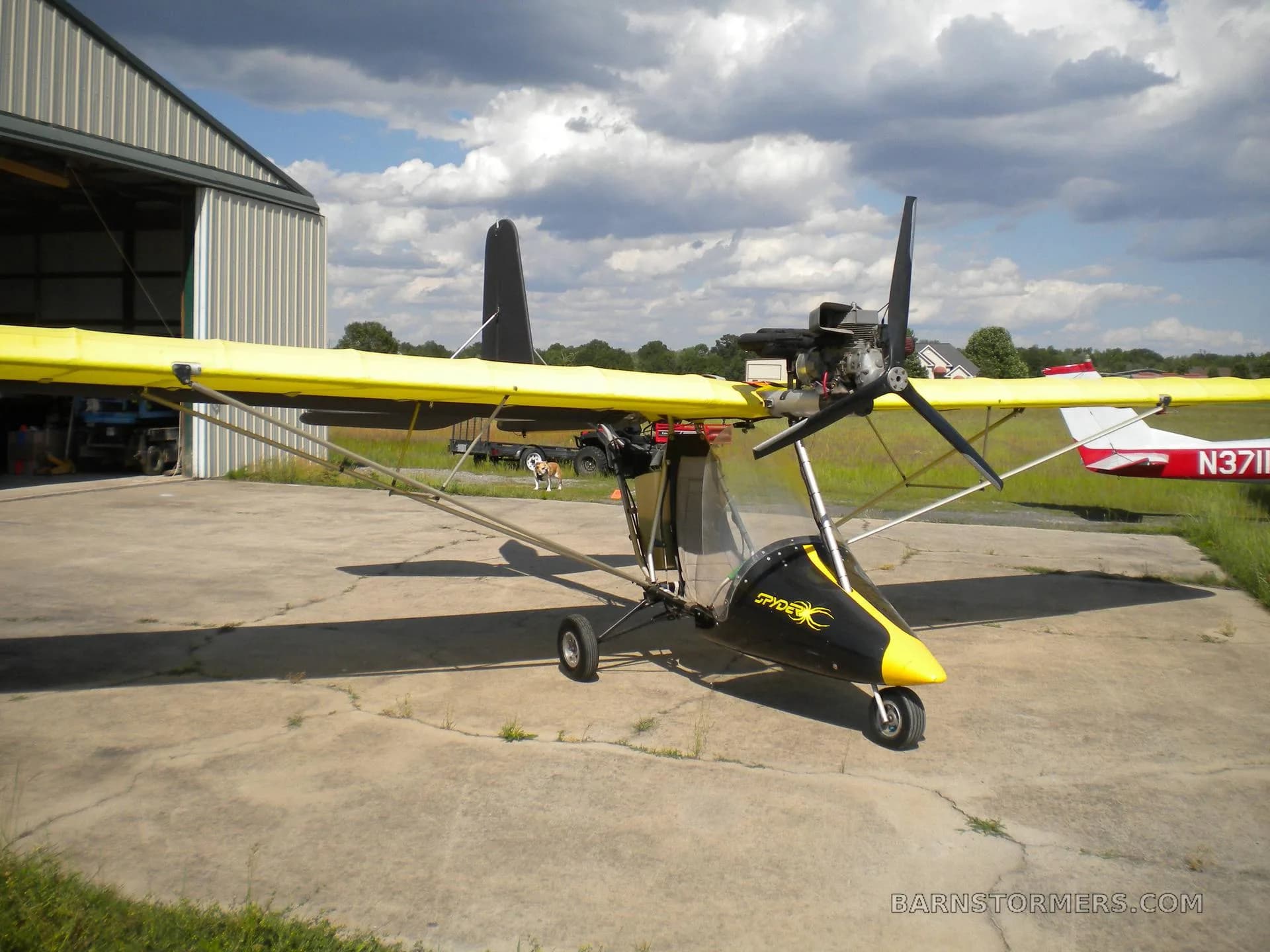 A yellow-winged ultralight aircraft with a black nose cone and 'Spyder' logo, parked in front of an open hangar.
