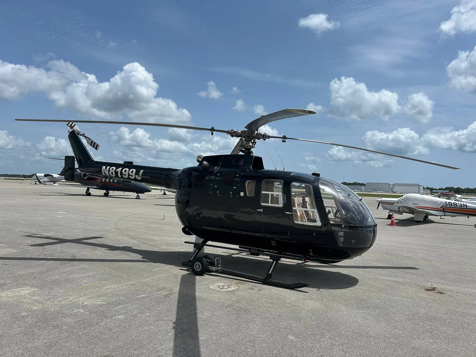 A black helicopter with tail number N8199J sits on the tarmac under a partly cloudy sky.